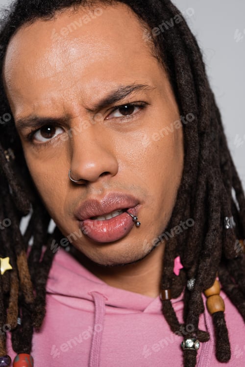 Preview: close up of pensive multiracial man in pink hoodie looking at camera isolated on gray