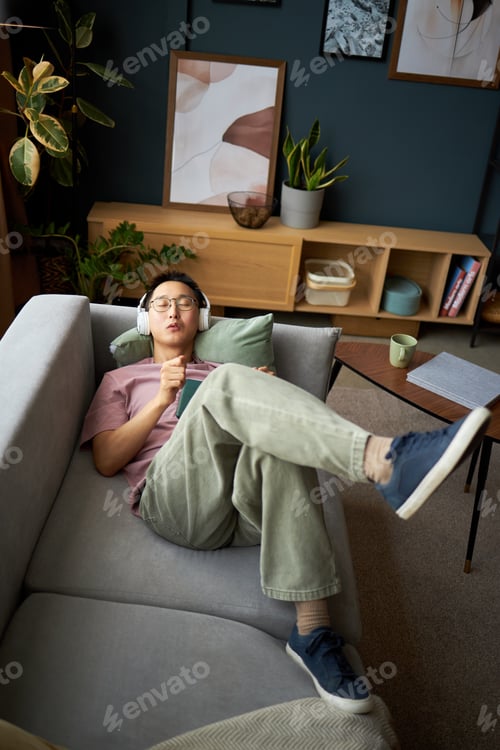 Preview: Young Adult Asian Man Listening to Music on Headphones While Relaxing on Couch