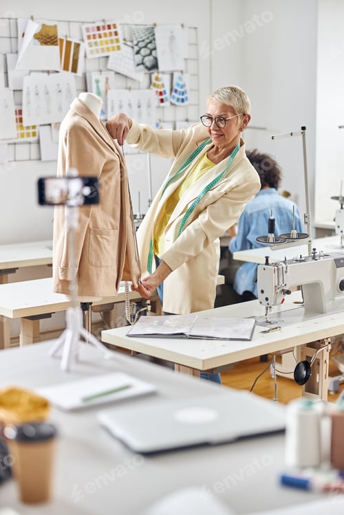 Preview: Positive middle aged female dressmaker presents new beige jacket shooting video