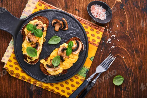 Preview: Scrambled eggs with fried mushrooms and basil on bread on black table background. Homemade breakfast