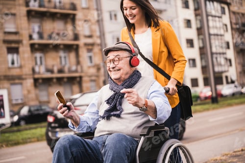 Preview: Laughing young woman pushing happy senior man with headphones and smartphone in wheelchair