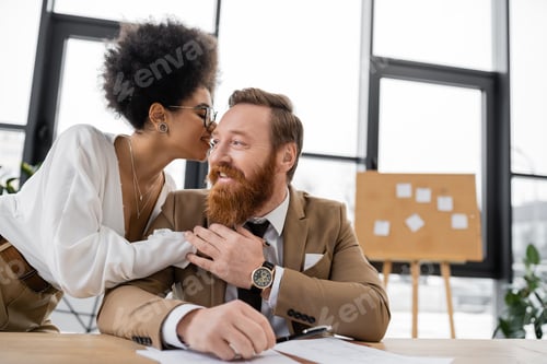 Preview: smiling african american businesswoman hugging while whispering in ear of cheerful coworker in