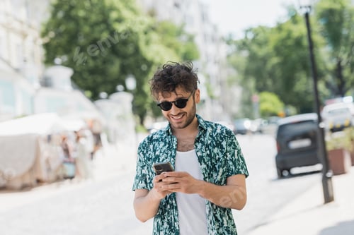 Preview: Overjoyed man in sunglasses walking and using smartphone on urban street