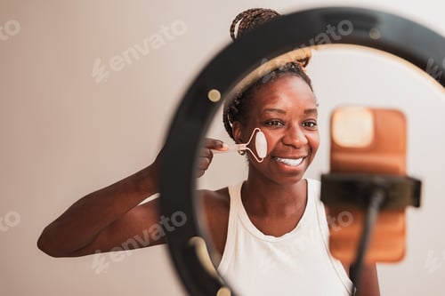 Preview: female blogger making a skincare video massaging face with jade roller in front of ring light