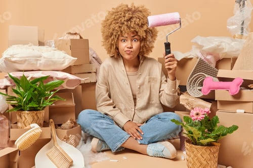 Preview: Serious curly haired woman holds paint roller busy doing redecoration of new house relocates to apar