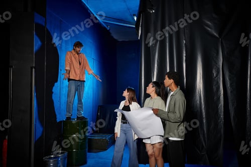 Vista previa: hombre pelirrojo mostrando un gesto de encogimiento de hombros y mirando a amigos multiculturales con un mapa en la sala de escape