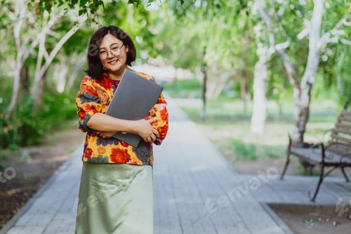 Preview: A fashionable, smiling Asian woman hugging a laptop is walking in the park