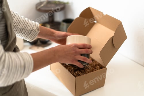 Preview: Female ceramist carefully packaging handmade mug for shipping in cardboard box