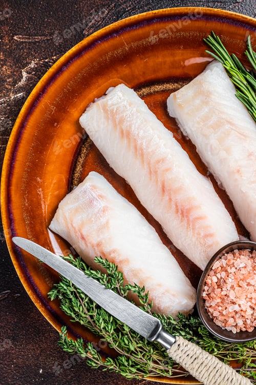 Preview: Raw Haddock loin fillet fish on rustic plate with thyme and salt. Dark background. Top view