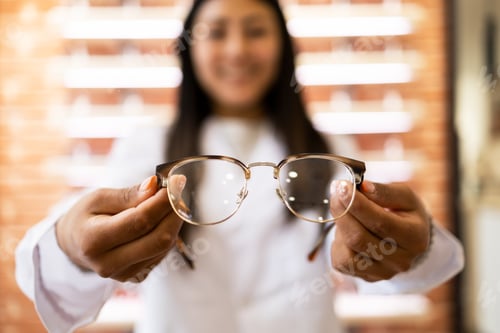 Preview: Blurred young eye doctor with long dark hair smiling and demonstrating optical glasses during work