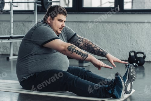 Preview: overweight tattooed man sitting on fitness mat and stretching at gym