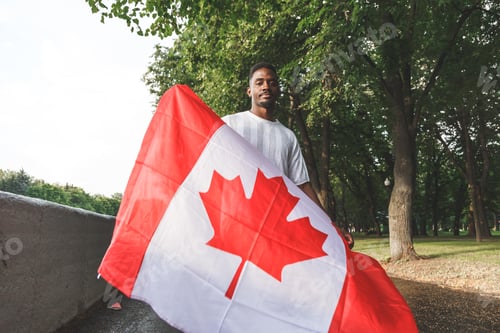 Preview: Handsome Afro American man with Canadian flag seriously looking at camera, standing outdoors.