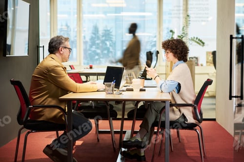 Preview: Side view of two coworkers sitting by table and using mobile gadgets