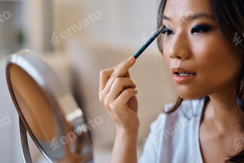 Preview: Young Asian woman applying eyeshadow while doing makeup at home.