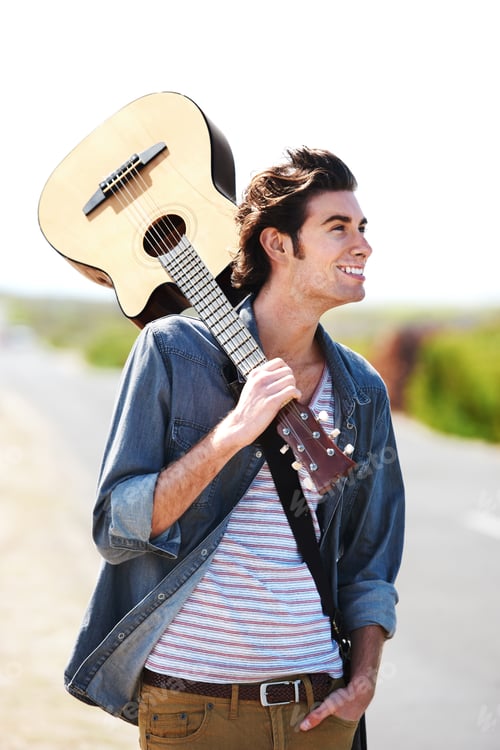 Preview: Smiling young man holding his guitar over his shoulder standing on the side of the road - closeup