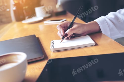 Preview: woman's hand writing on a blank notebook with laptop , tablet and coffee cup on wooden table