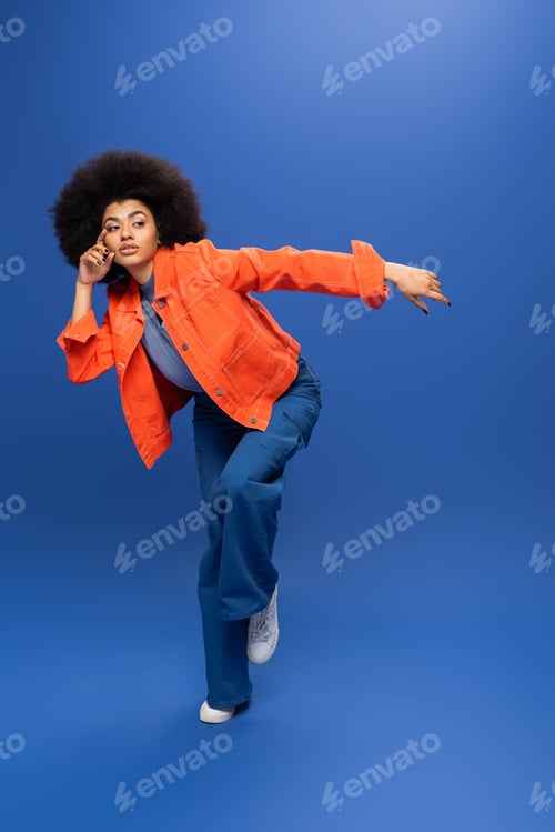 Preview: Full length of young african american woman in stylish outfit looking away on blue background