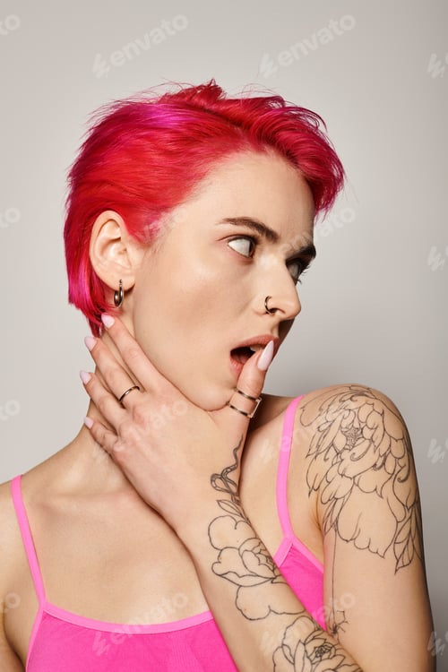 Preview: tattooed and pierced woman with pink hair and rings on her fingers looking away on grey background