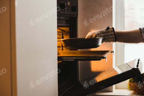 Preview: Man's hand in potholder puts mold with dish in the embedded oven in kitchen at home