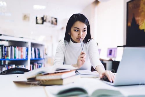 Preview: Elegant adult ethnic woman thinking while sitting at laptop and looking away in modern library