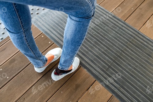 Preview: Female legs in denim jeans pants and light summer sneakers standing on wooden floow