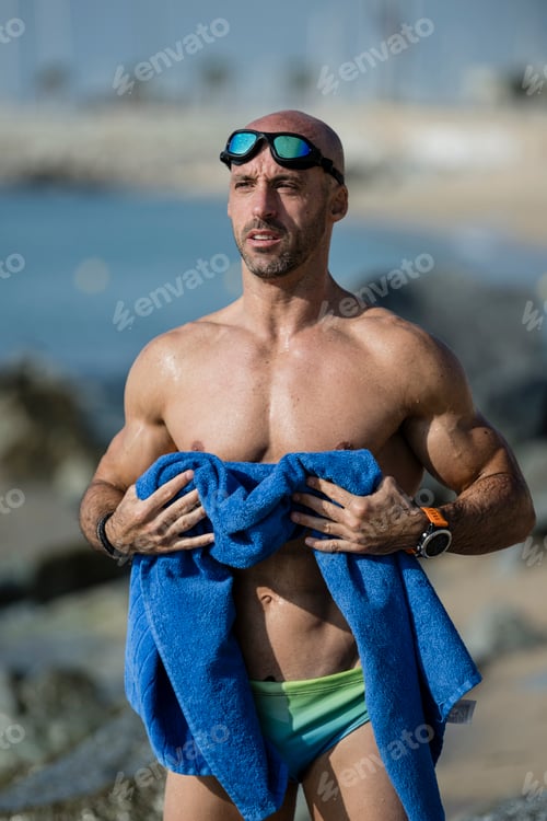 Preview: Sexy muscular swimmer toweling off his body with swim glasses at the beach. Fit man with a blue