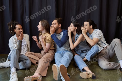 Preview: multicultural team sitting and telling secret to surprised colleague near black drape in office