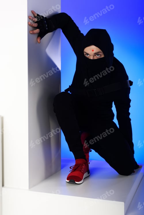 Preview: ninja in black clothing with katana behind standing on white block isolated on blue