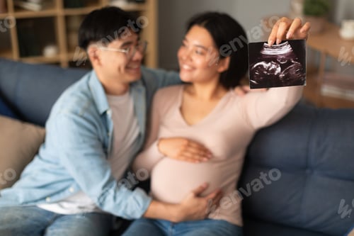 Preview: Happy pregnant asian couple showing ultrasound image at home