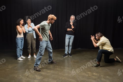 Preview: young actors playing scene near grey haired art director and amazed interracial women. Translation