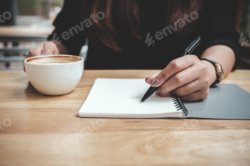 Preview: a woman's hand writing down on a white blank notebook while drinking coffee