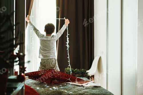 Preview: Boy decorating home and window for Christmas with garland and red star Magic winter holiday spirit