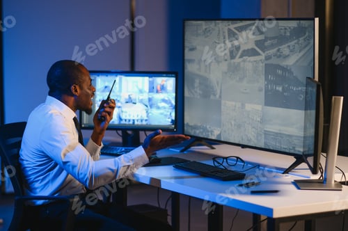 Preview: African American security guard looking at desktop computer screen while speaking on walkie talkie