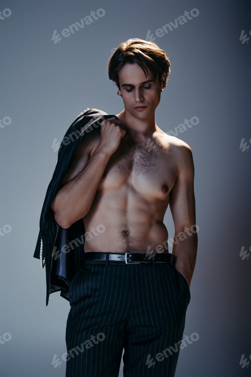 Preview: shirtless man holding blazer and posing with hand in pocket of trousers on grey