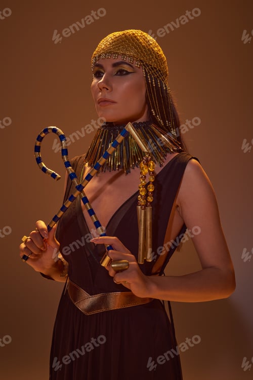Preview: Elegant woman in egyptian golden headdress and look holding crook and flail on brown background