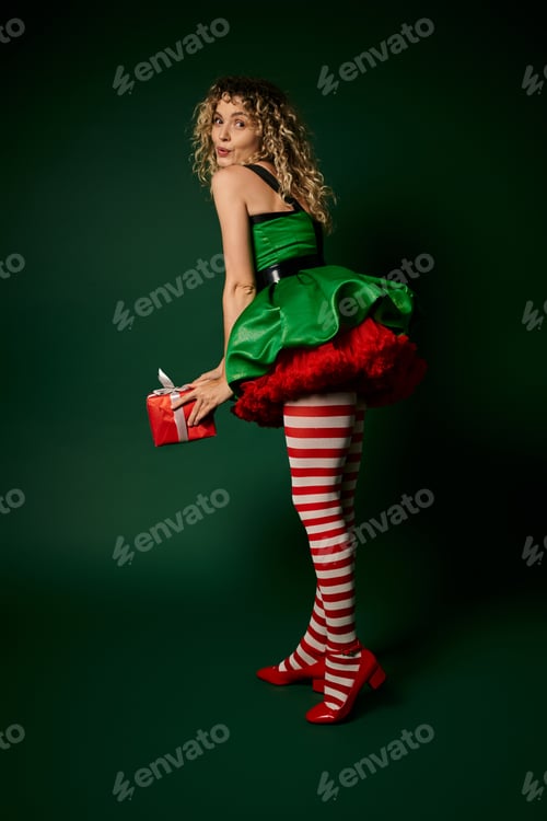 Preview: jolly woman in new year elf costume posing with pouted lips and holding huge present in hands