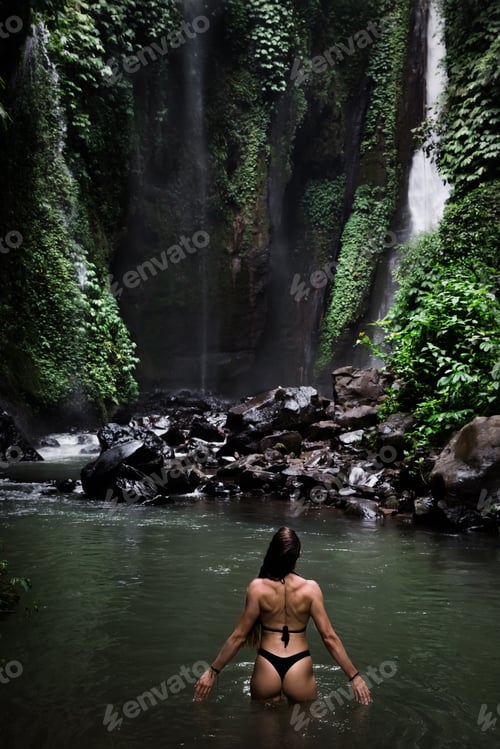 Preview: Back view of beautiful woman bathing and relaxing in natural swimming pool before waterfall in