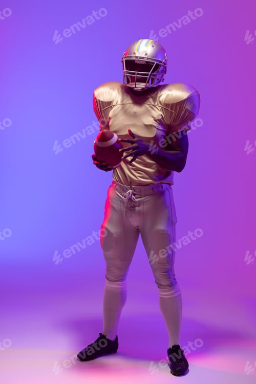 Preview: African american male american football player holding ball with neon blue and purple lighting