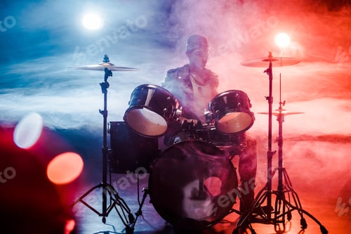 Preview: male musician in leather jacket playing drums during rock concert on stage with smoke and spotlights