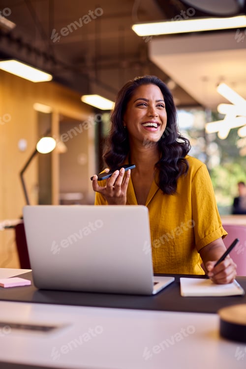 Preview: Mature Businesswoman Working On Laptop At Desk In Office Talking Into Mic Of Mobile Phone