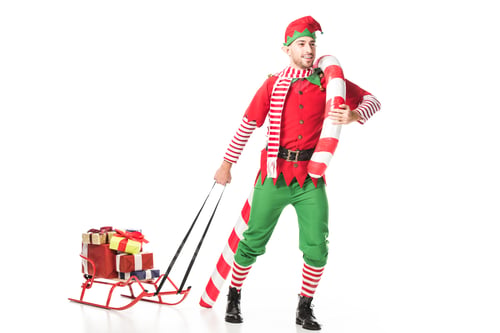 Preview: happy man in christmas elf costume carrying sleigh with presents and big candy cane isolated on