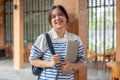 Preview: Happy asian woman holding bag strap carrying laptop while standing walking by wooden pillar in cafe.