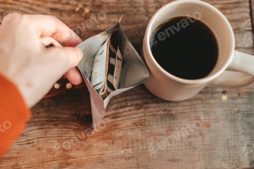 Preview: hand opening a package with disposable drip coffee, next to a cup, on a wooden background.