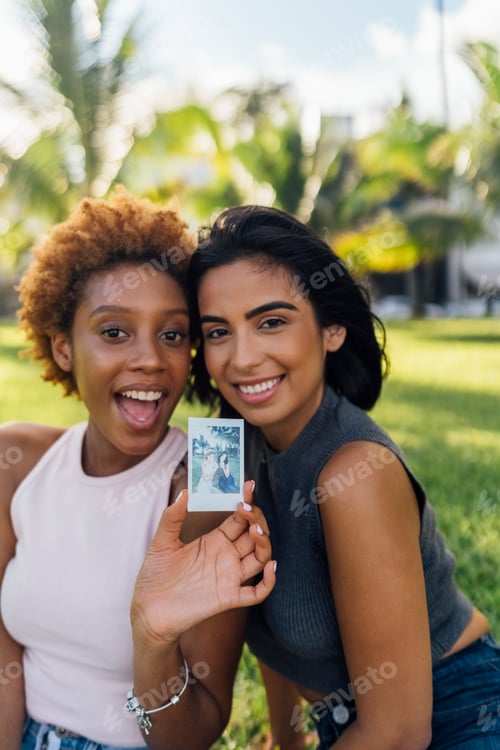 Preview: Two happy female friends showing an instant photo in a park