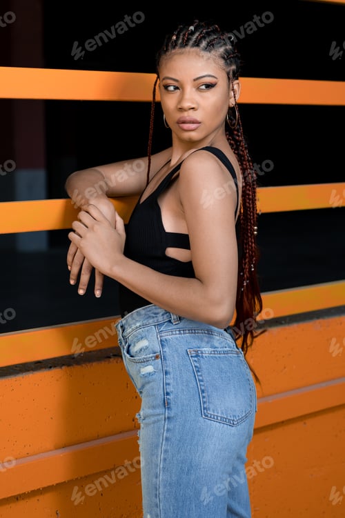 Preview: Vertical shot of a cool young Afro-American lady with deads leaning on an orange construction