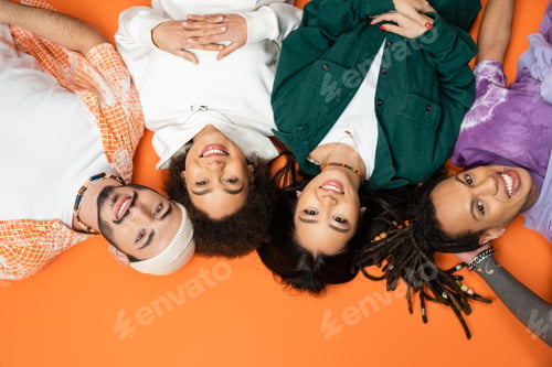 Preview: top view of pleased and trendy multiethnic friends lying and smiling at camera on orange background