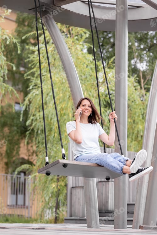 Preview: caucasian woman in stylish clothes riding on a swing and talking on the phone and smiling