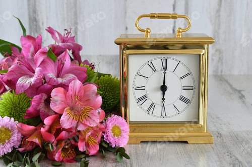 Preview: Classic timepiece, gold clock with roman numerals setting next to a bouquet of pink & green flowers