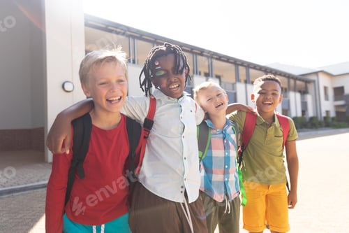 Preview: Happy multiracial elementary schoolboys with arm around standing at school campus during sunny day