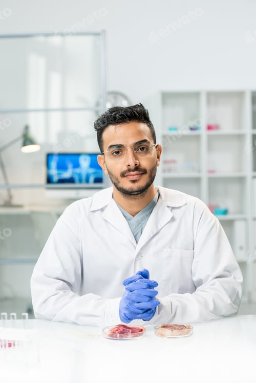 Preview: Young gloved male scientific researcher in whitecoat looking at you by workplace